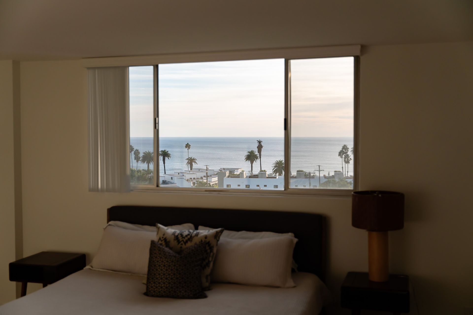 Bedroom with bed and window overlooking the ocean, palm trees, and buildings.