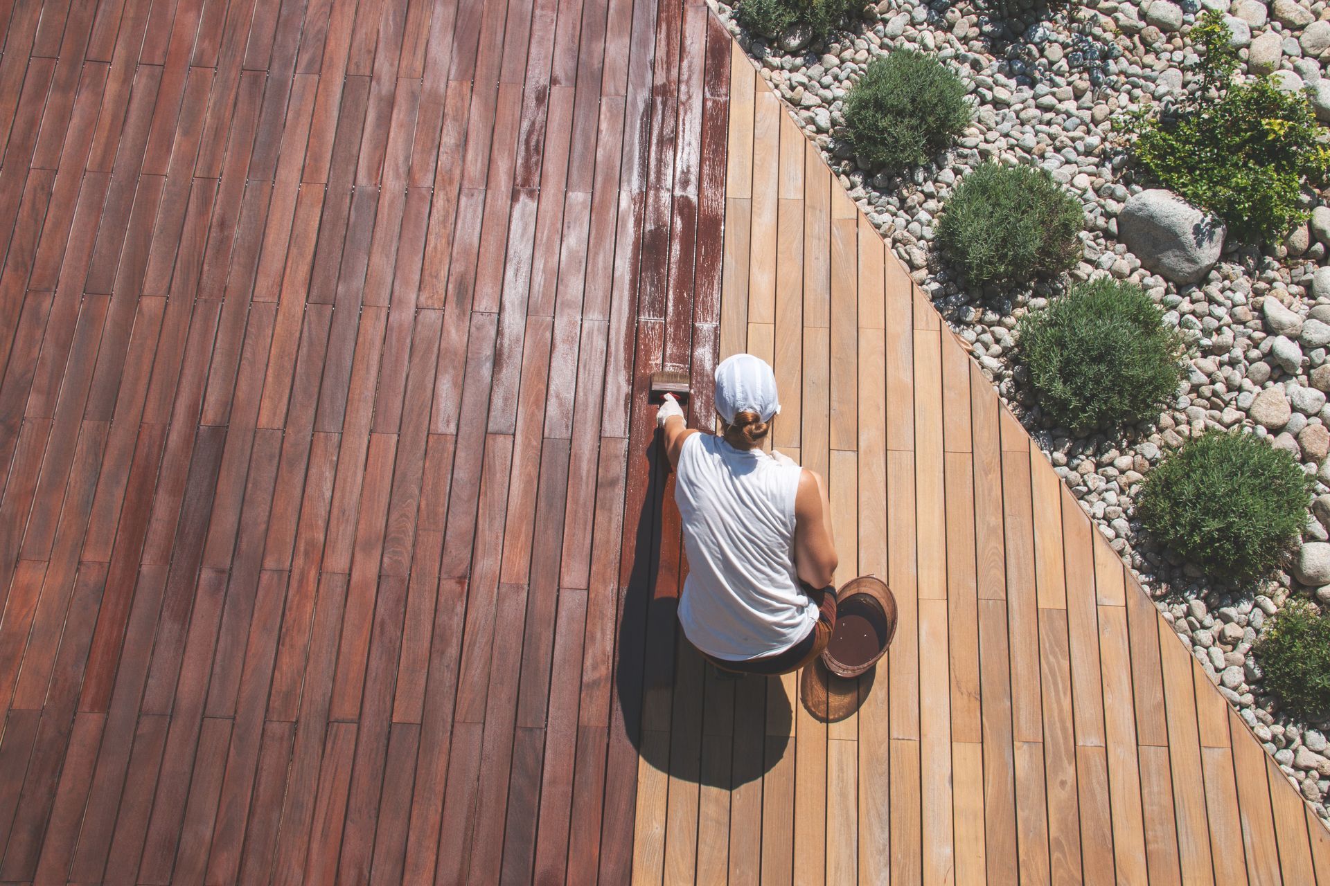 Overhead view of a person meticulously applying protective wood stain with a brush during the renovation treatment of a wood deck.