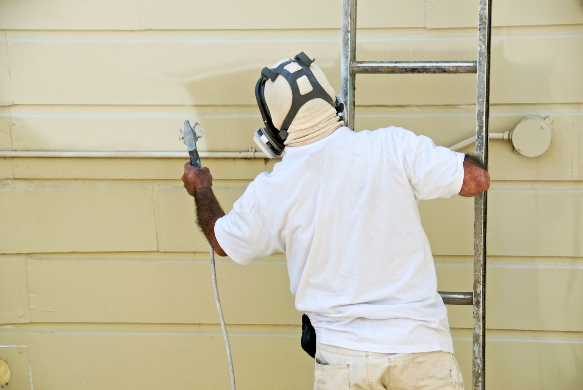 A skilled handyman on a ladder expertly operates a paint spray gun, applying a smooth coat of paint to the exterior surface.