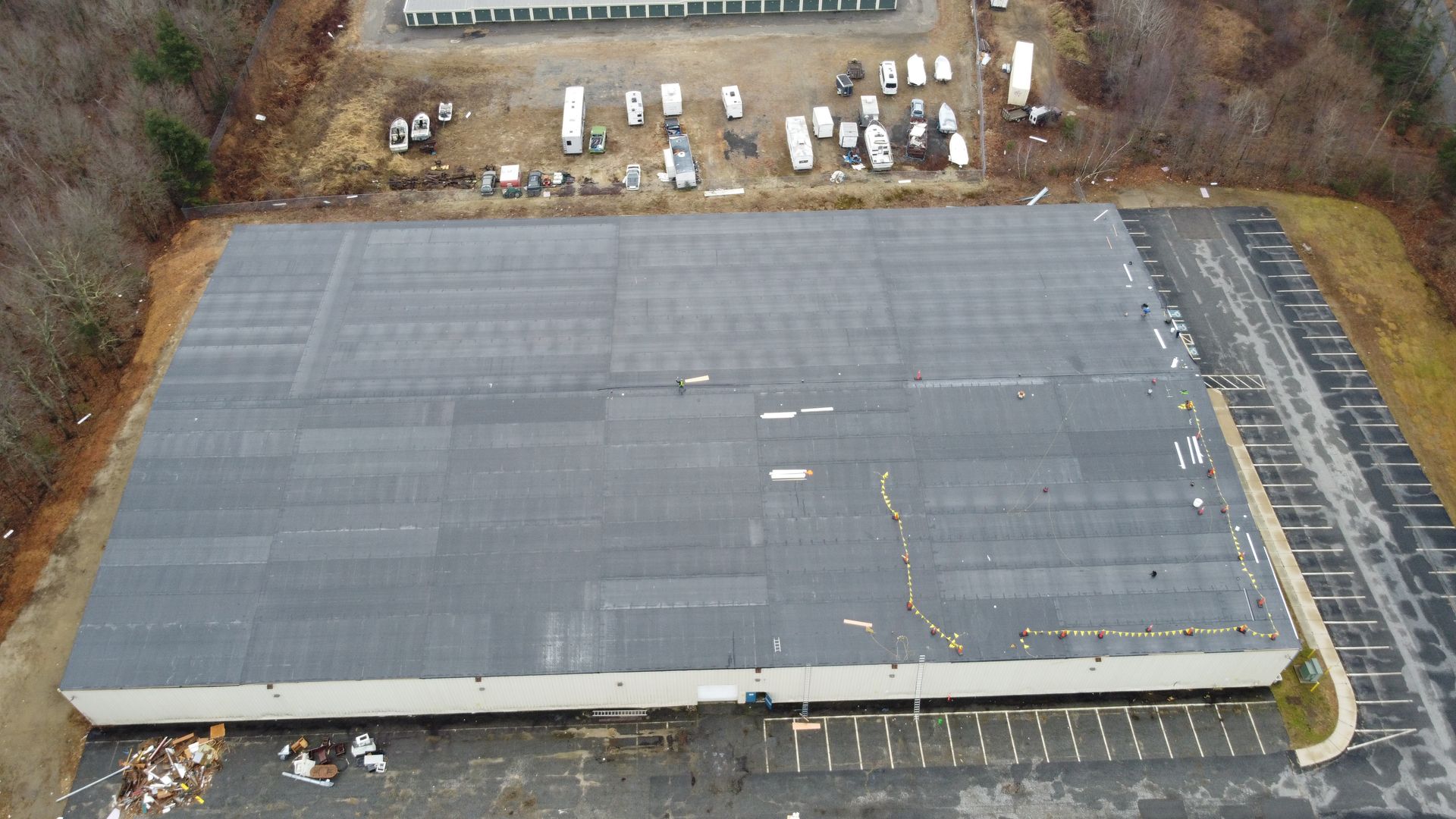 Overhead view of a large industrial building with a flat roof and parking lot. Surrounding trees and a storage facility.