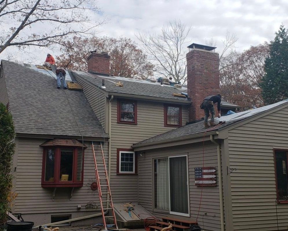 Roofers working on a house with a chimney and American flag. Overcast day, brown siding, gray roof.