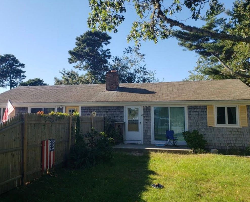 Ranch-style house with gray shingles and yellow window shutters; American flag on wooden fence. Sunny day.