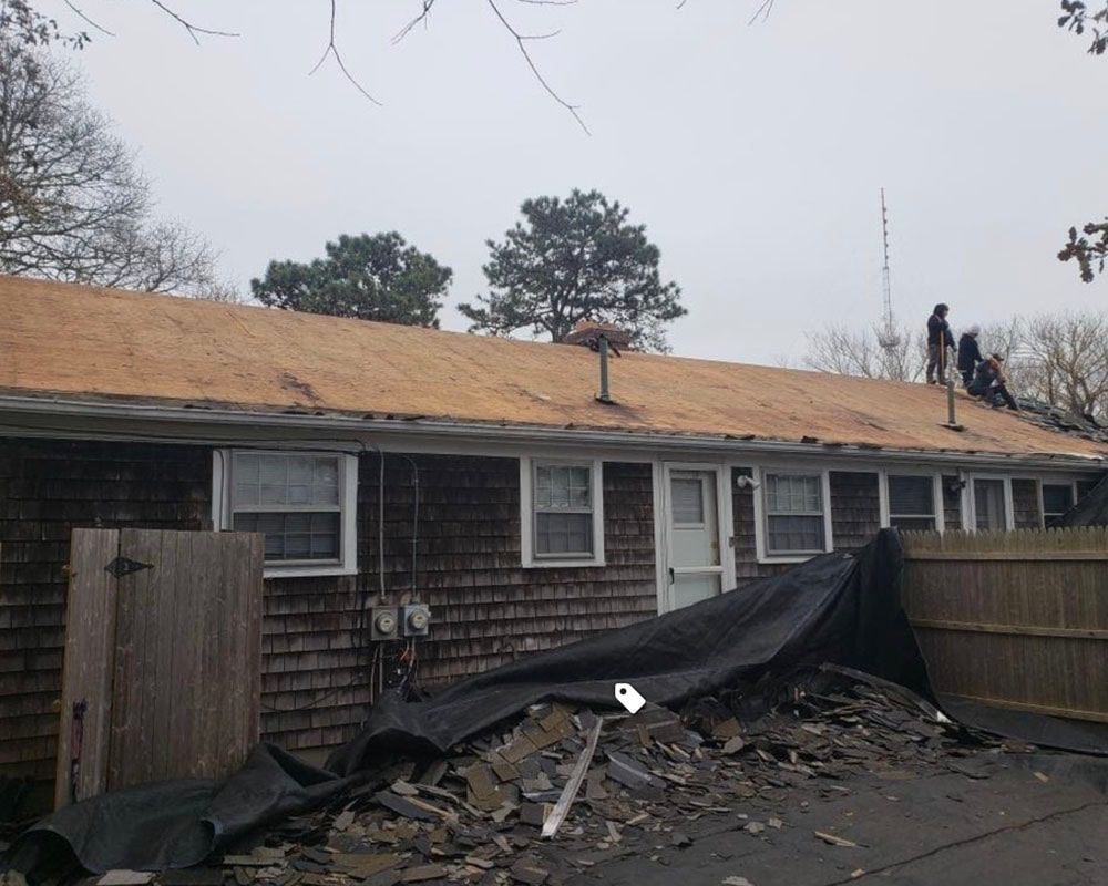 Roofers removing old shingles from a residential building on a cloudy day. Debris is covered by a tarp.