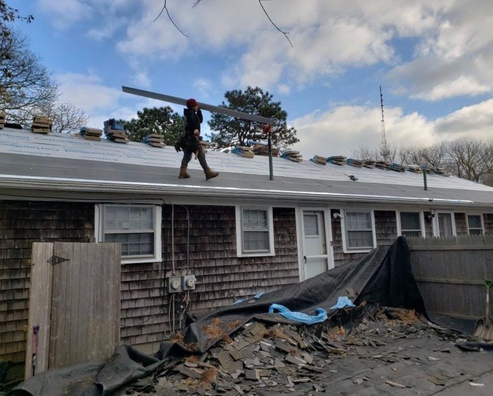 Roofer carries a metal beam across a roof; a pile of shingles and debris in the foreground.
