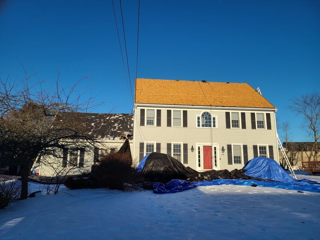 Two-story house with new roof partially installed; snow on ground, blue tarps, and a bright blue sky.
