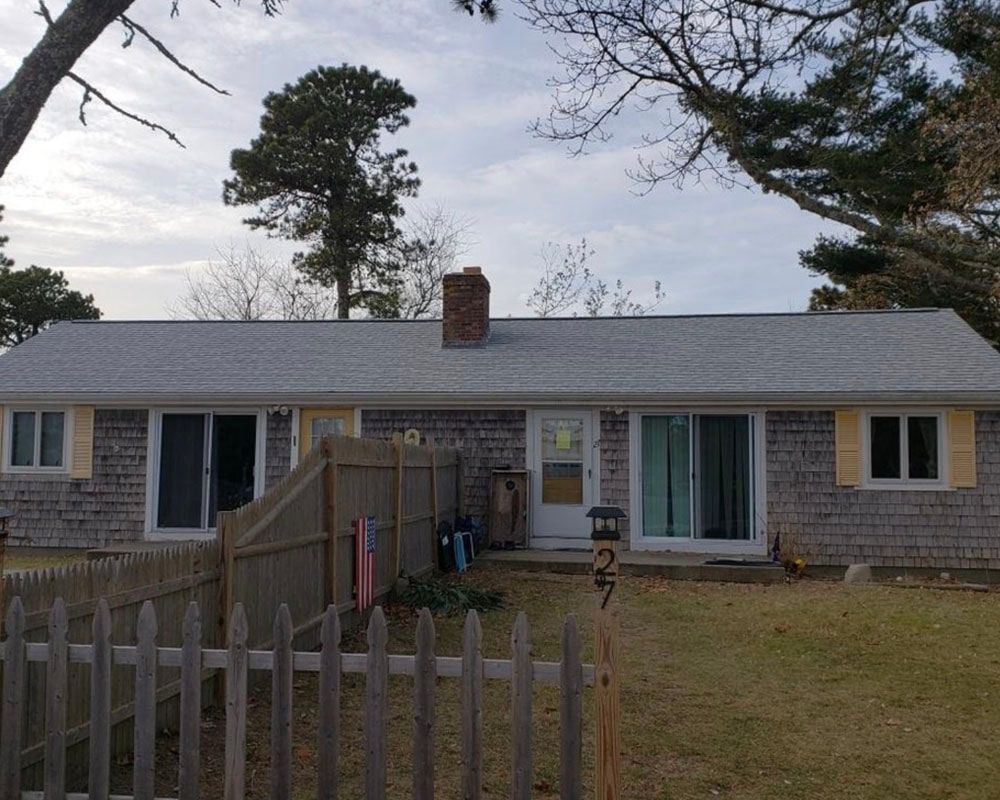 Ranch-style house with gray roof and cedar shingle siding, number 27, with a wooden fence in front.