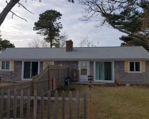 A small, weathered house with a gray roof, wooden shingles, and a picket fence in front.