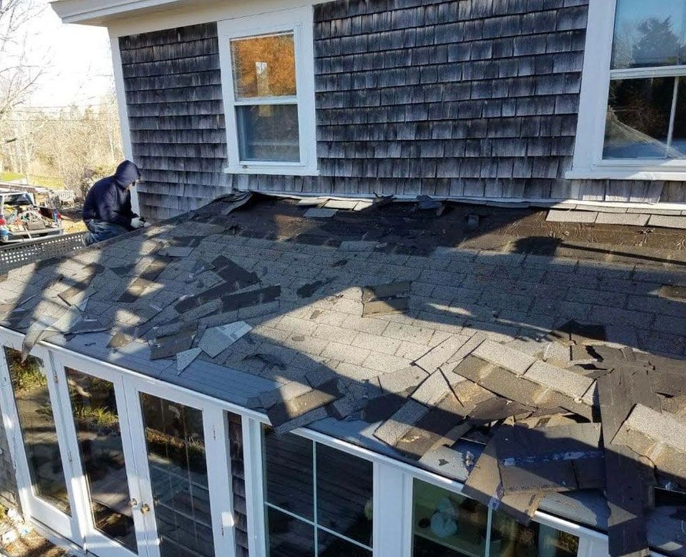Person on a house roof removing old shingles. Weathered wood siding, windows, and sunlight.