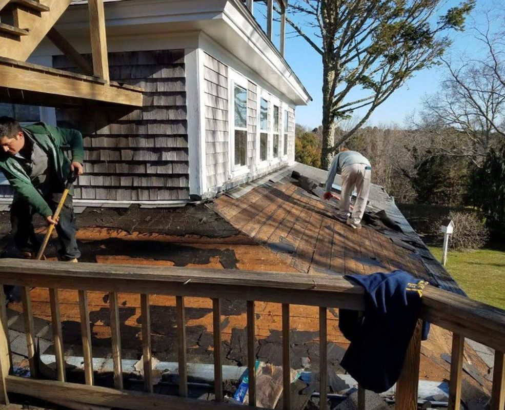 Two workers removing roof shingles from a house with wood shingle siding and a wooden deck.
