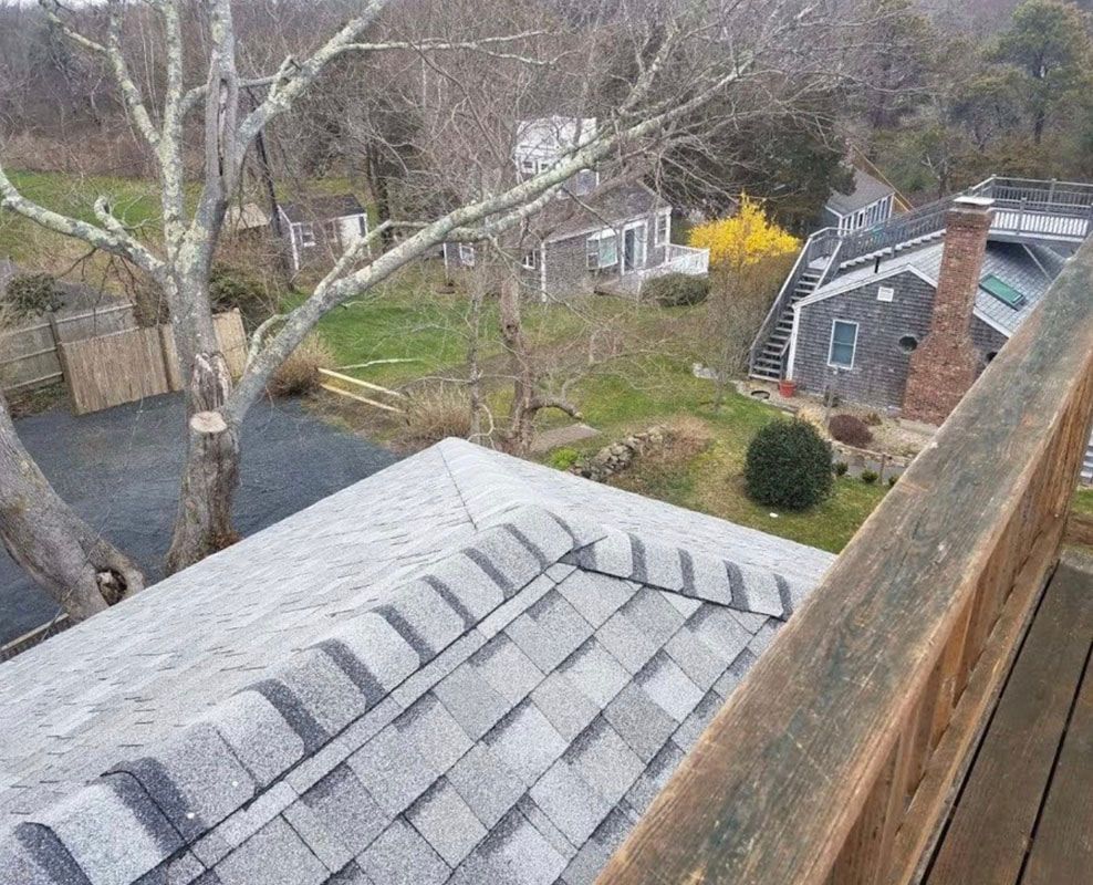 View from a deck: gray shingled roof in foreground, houses and trees in background on a cloudy day.