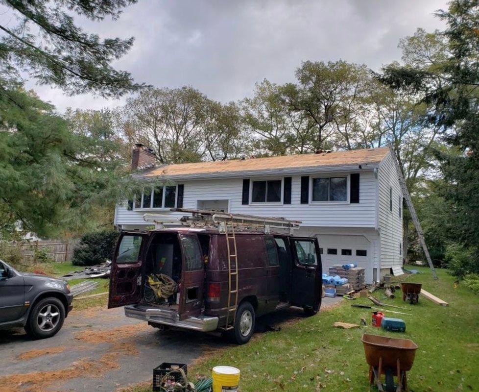 A brown van parked in front of a two-story white house with a roof being worked on.