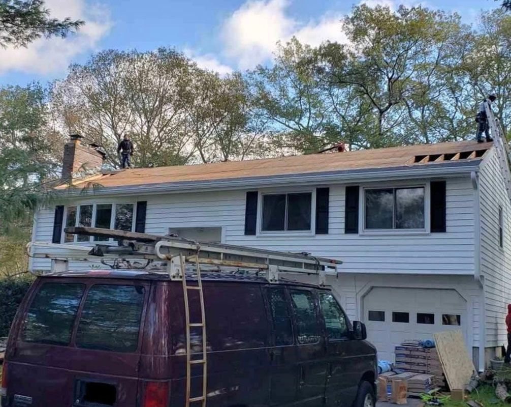 Roofers working on a two-story white house; van and ladder in foreground.