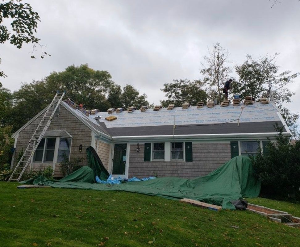 House roof under construction with a worker; green tarp on the grass and a ladder. Cloudy day.