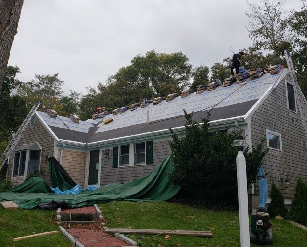 Roofers replacing shingles on a house; gray siding, green tarp, cloudy sky.