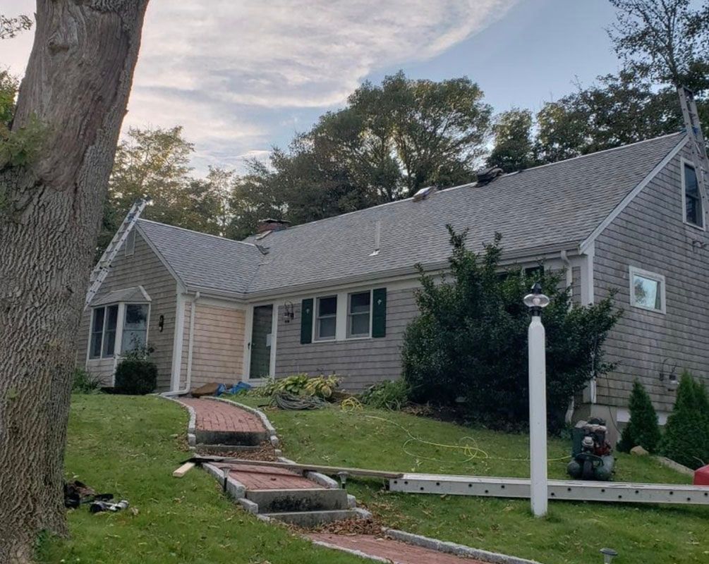 A gray-shingled house with a brick walkway on a grassy lawn, under a cloudy sky.