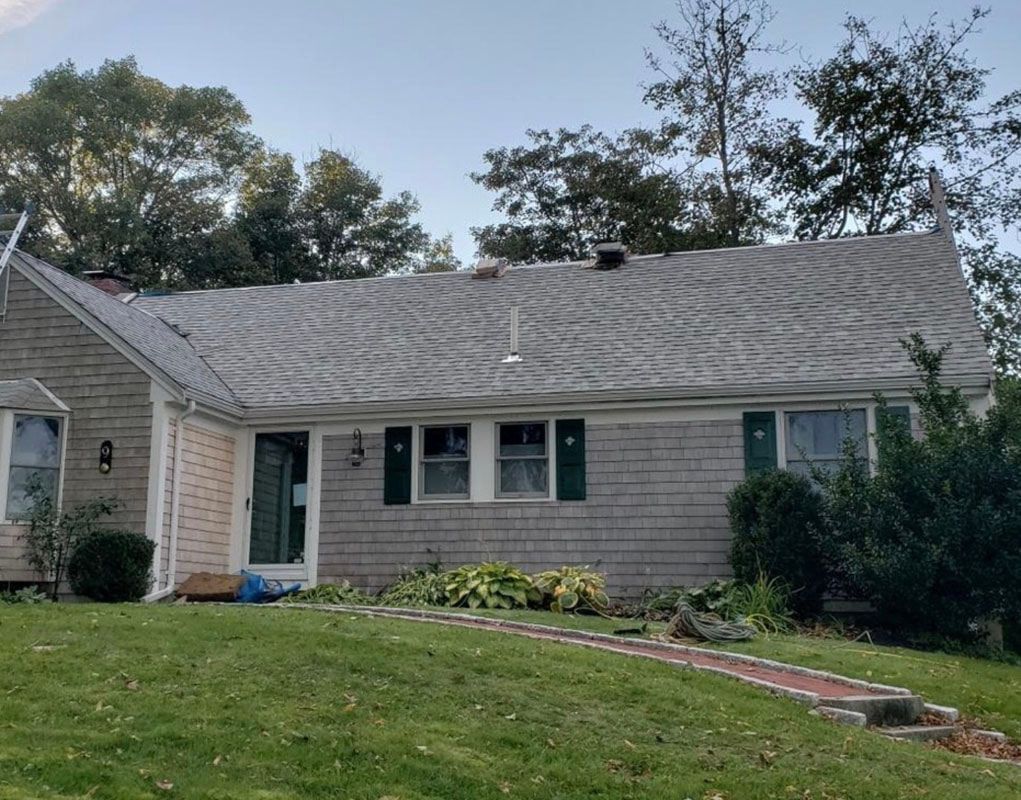 A one-story house with a gray shingle roof and green shutters on a lawn.