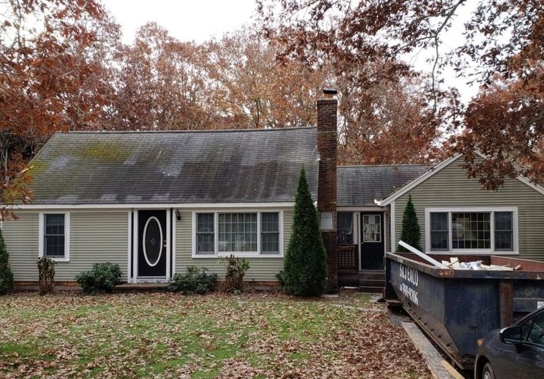 Beige house with visible moss on roof and a dumpster in the driveway. Brown trees in the background.