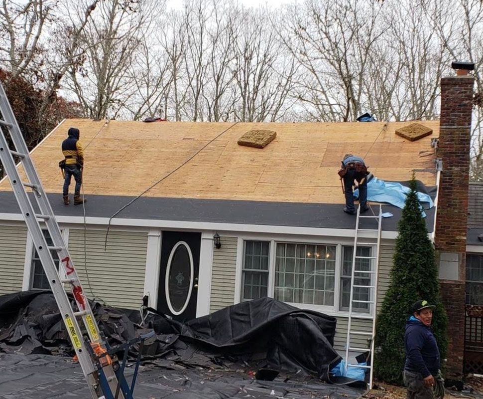 Roofers installing a new roof on a light green house. Ladders, tarps, and a brick chimney visible.