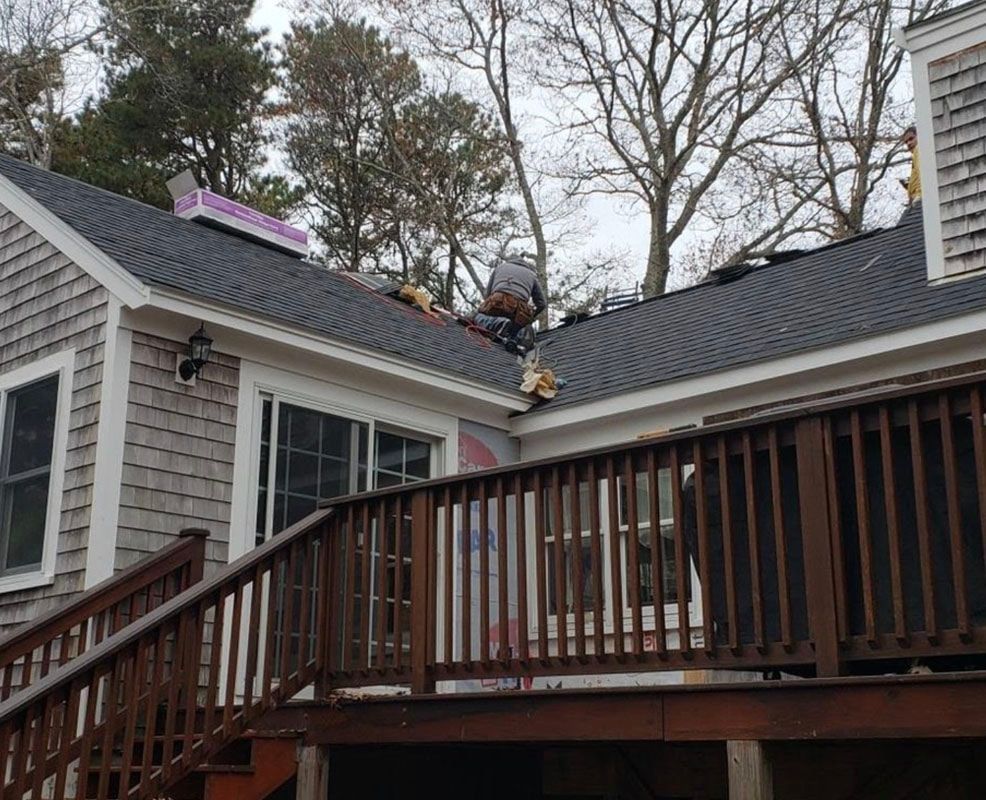 Roofers working on a dark shingle roof of a light gray house with a wooden deck.