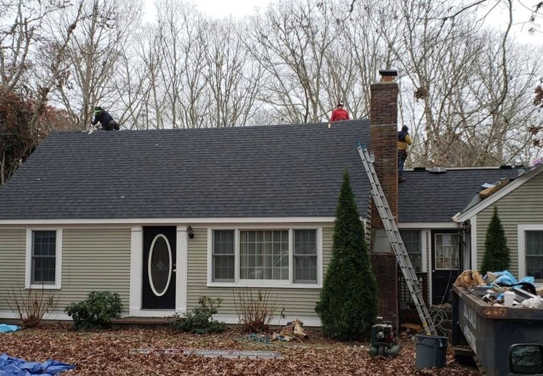 Roofers working on a dark gray shingle roof of a beige house with a black door.