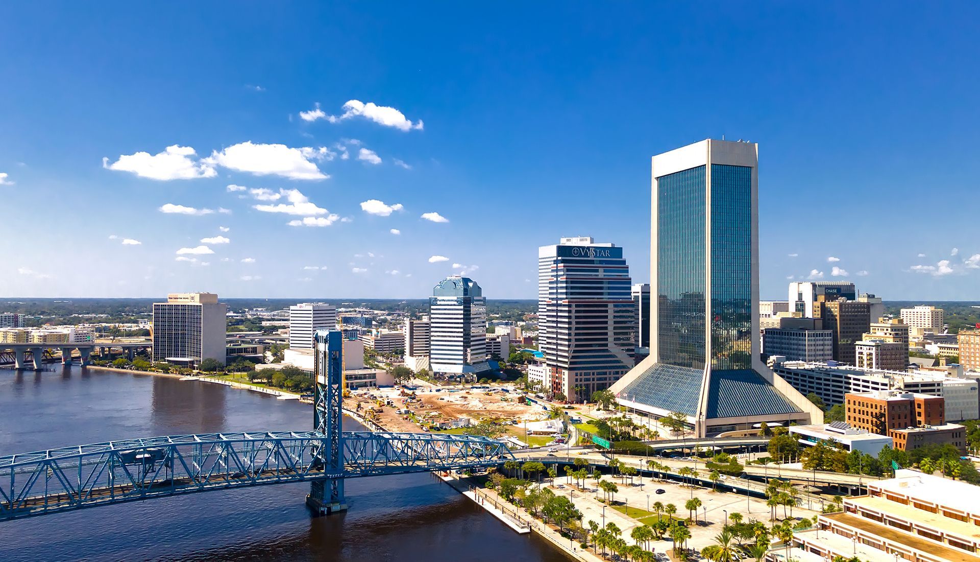 Skyline of Jacksonville, Florida, with buildings along the water. Blue bridge in foreground, blue sky with clouds.