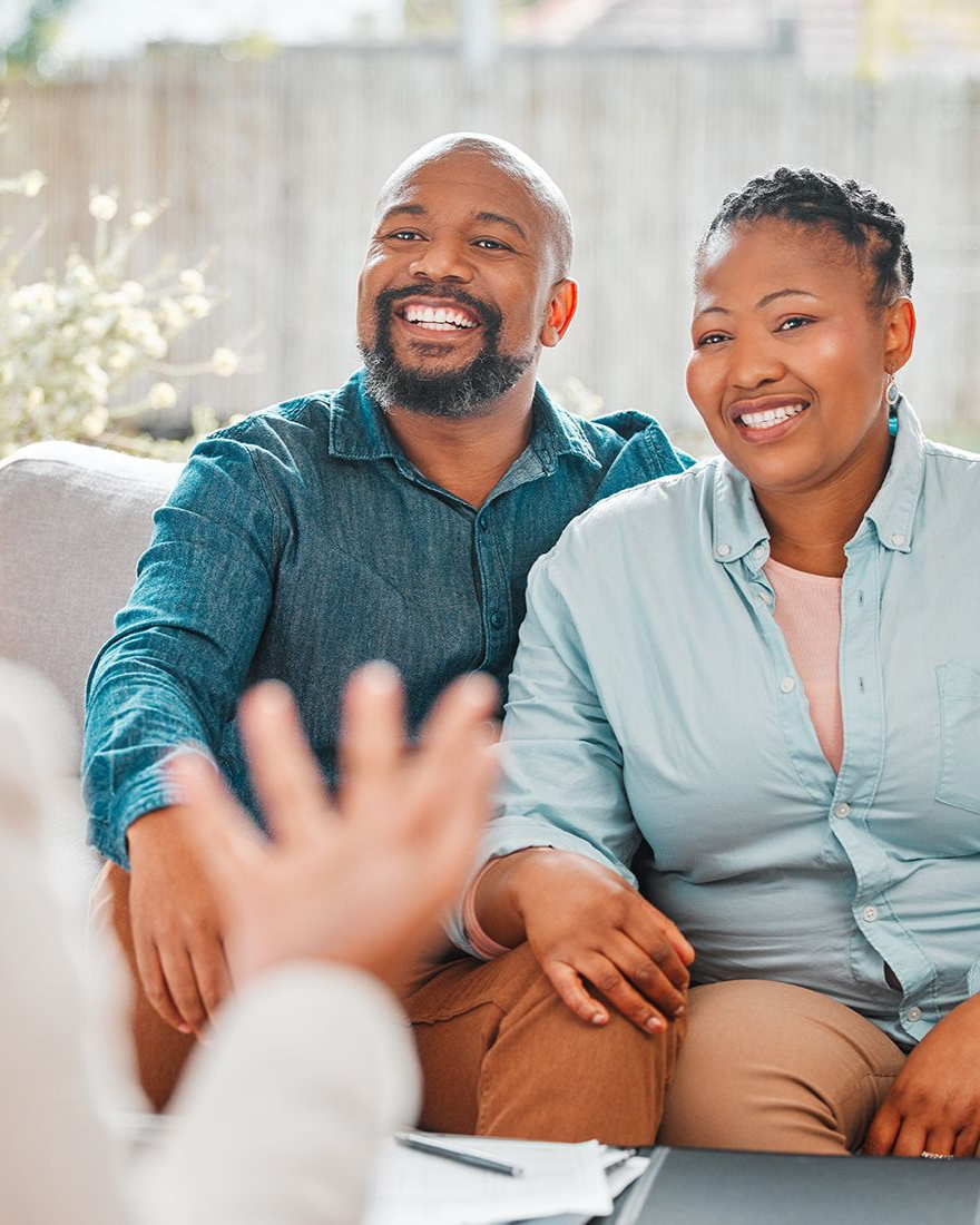 Couple smiles while listening, someone's hand raised in front of them, documents visible.