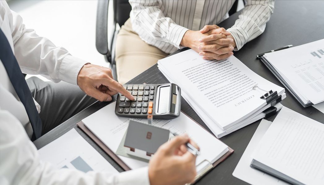 Two people at a desk review documents and use a calculator; a person points to the calculator.