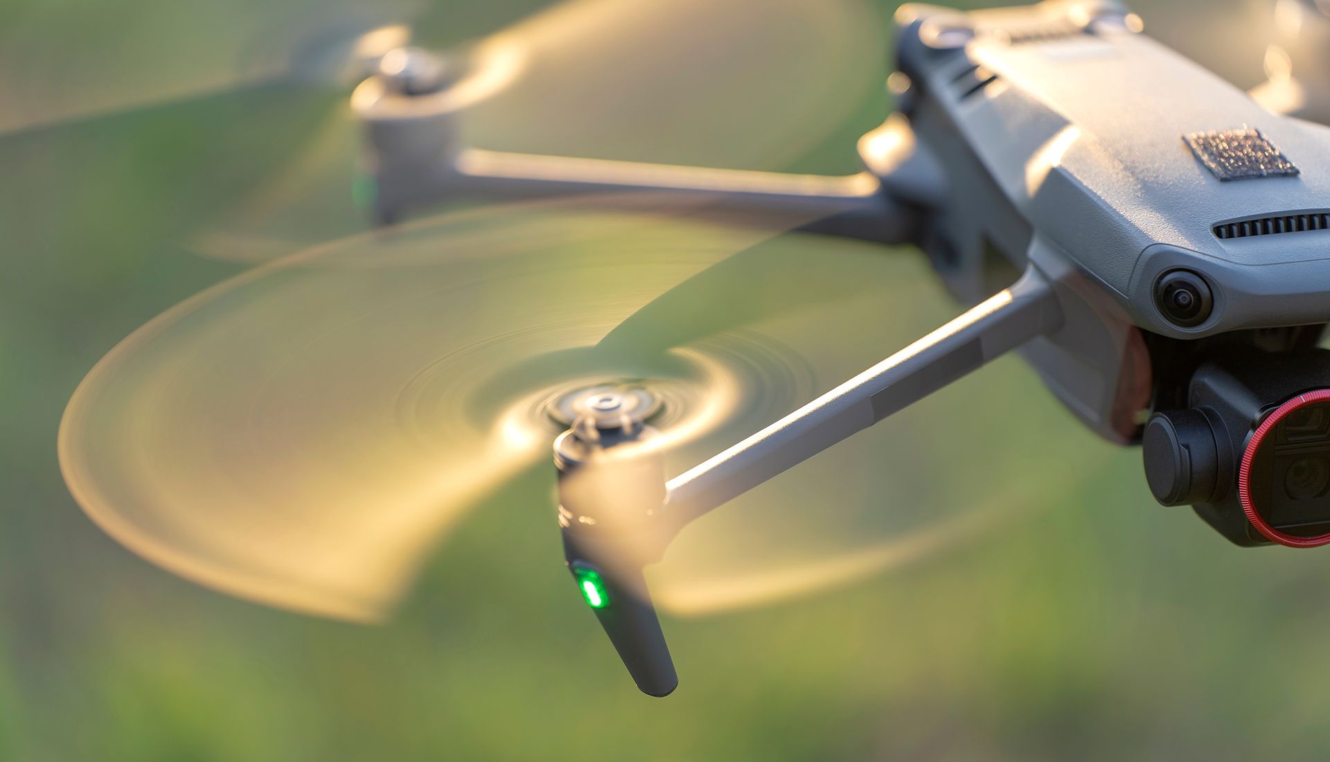 Drone with spinning propellers, partly lit by sunlight against a blurred green background.