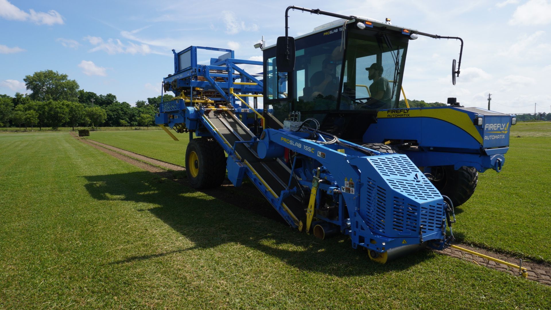 Blue agricultural harvester in a grassy field under a clear sky, shown from the front-left side.
