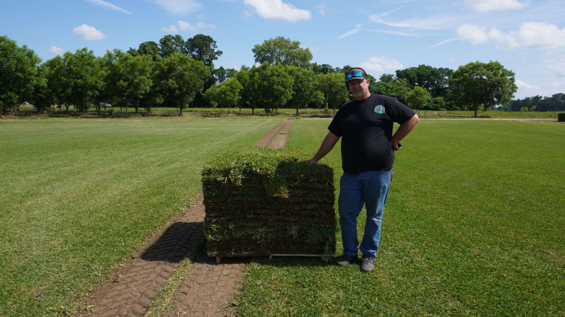 Man stands by a stack of sod on a cart in a field with trees in the background.