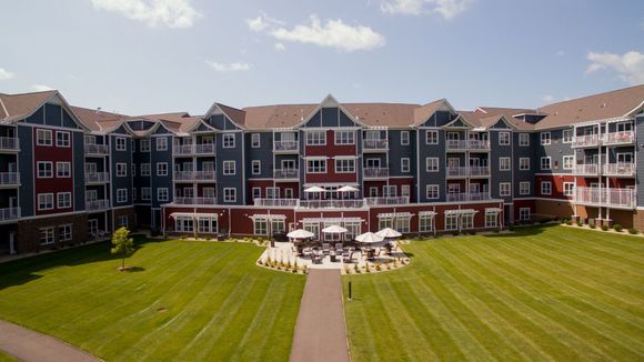 Multi-story apartment building with red and blue siding, featuring a central courtyard with a patio, umbrellas, and lawn.