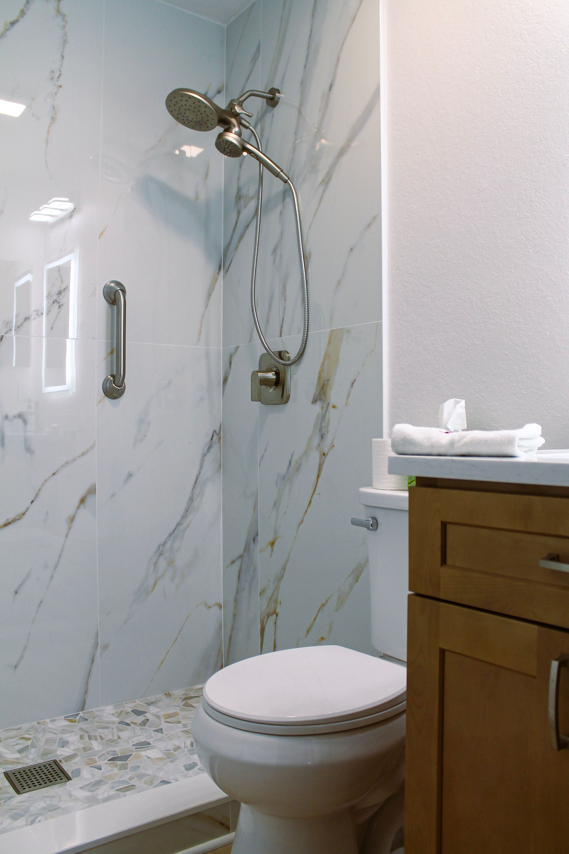 Modern white marble look large form tile shower with modern brushed stainless steel spa style showerhead with brushed silver accessibility handle and pebble mosaic flooring, brushed stainless steel drain, white modern toilet, Honey oak colored cabinets with modern brushed stainless pull