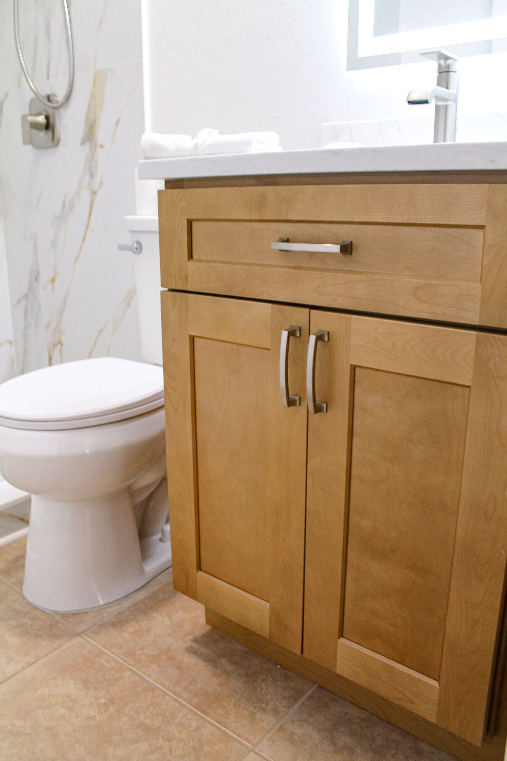 Modern bathroom with wood honey oak cabinets with modern brushed stainless steel pull, white toilet and modern brushed stainless steel tap on quartz countertop, white tile shower coral colored tile flooring