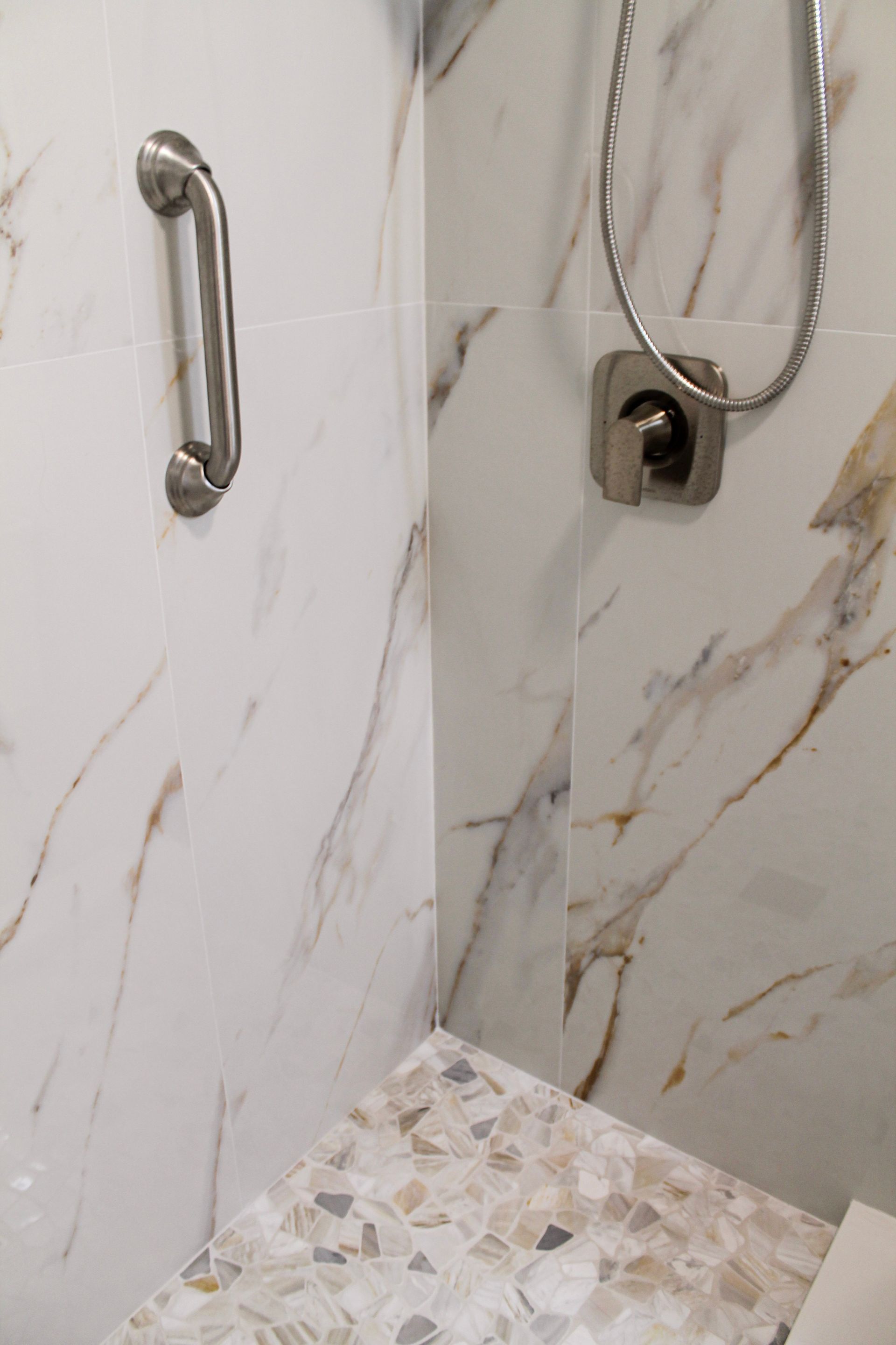 Modern white marble look large form tile shower with modern brushed stainless steel spa style showerhead with brushed silver accessibility handle and pebble mosaic flooring, brushed stainless steel drain, white modern toilet