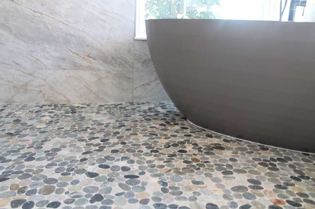 Grey bathtub with large form tile wall and pebble mosaic flooring with large window and natural lighting