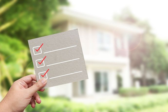 Hand holding a card with a checklist, three items checked, in front of a house.