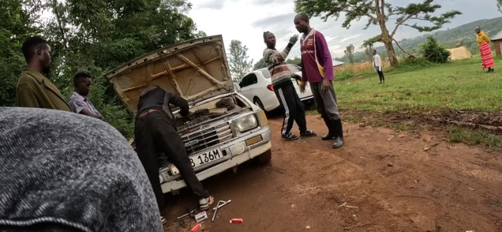 People gather around a car with an open hood on a dirt road, possibly working on the engine.