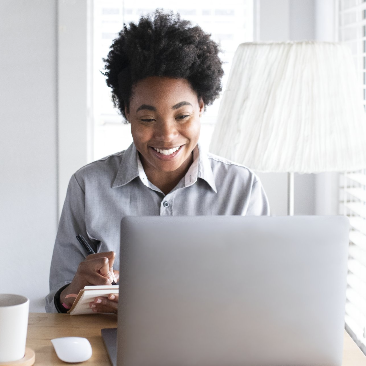 Woman smiling, taking notes while using a laptop, sitting at a desk by a window.