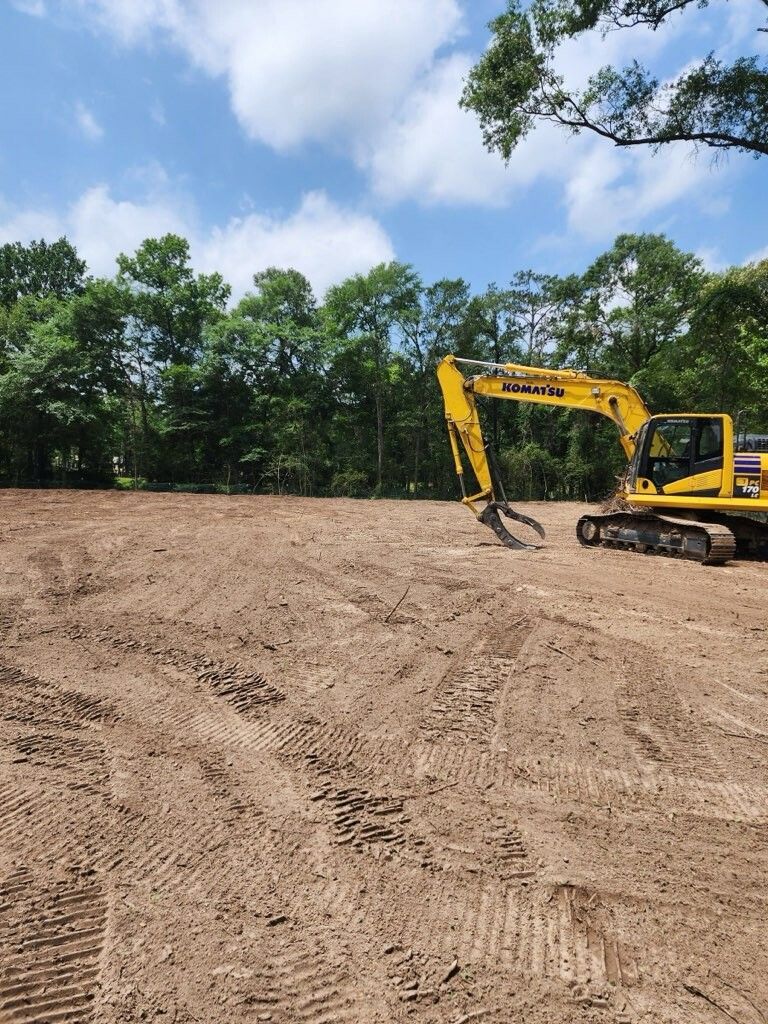 A yellow excavator on a leveled dirt lot with trees in the background under a partly cloudy sky.