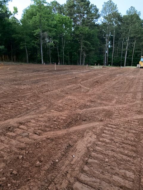 Cleared land with dirt rows, stakes, and tire tracks. Trees in the background.