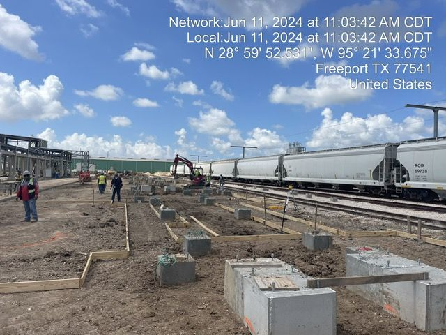 Construction site in Freeport, TX, with workers, machinery, and a passing train on a sunny day.