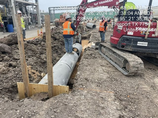 Construction workers installing pipe in a trench near Freeport, TX. A backhoe and wooden supports are visible.