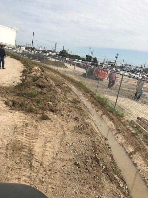 Dirt path and ditch next to a chain link fence and a large parking area with vehicles. People are walking.