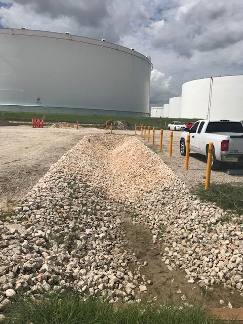 Gravel-lined trench near large white storage tanks. A white truck and posts mark a path. Cloudy sky.