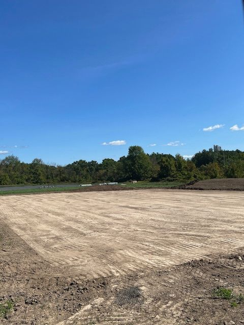 Cleared dirt lot with tractor tracks, blue sky, trees in the background.