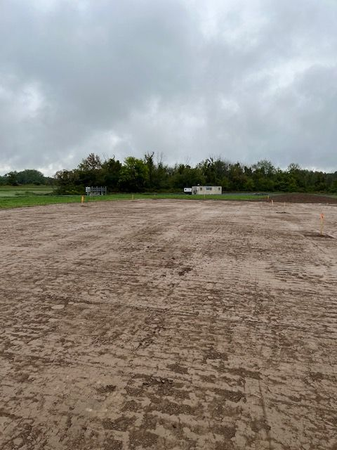 Muddy field with overcast sky, a few buildings, and trees in the distance.
