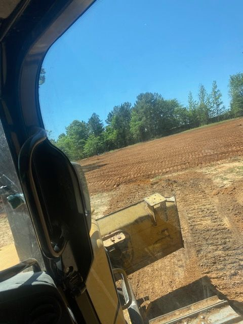 View from inside a bulldozer cab, overlooking a cleared field and trees under a blue sky.