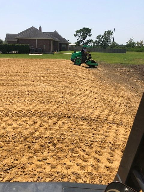 A person operating a green tractor tilling a large, sandy field near a house.