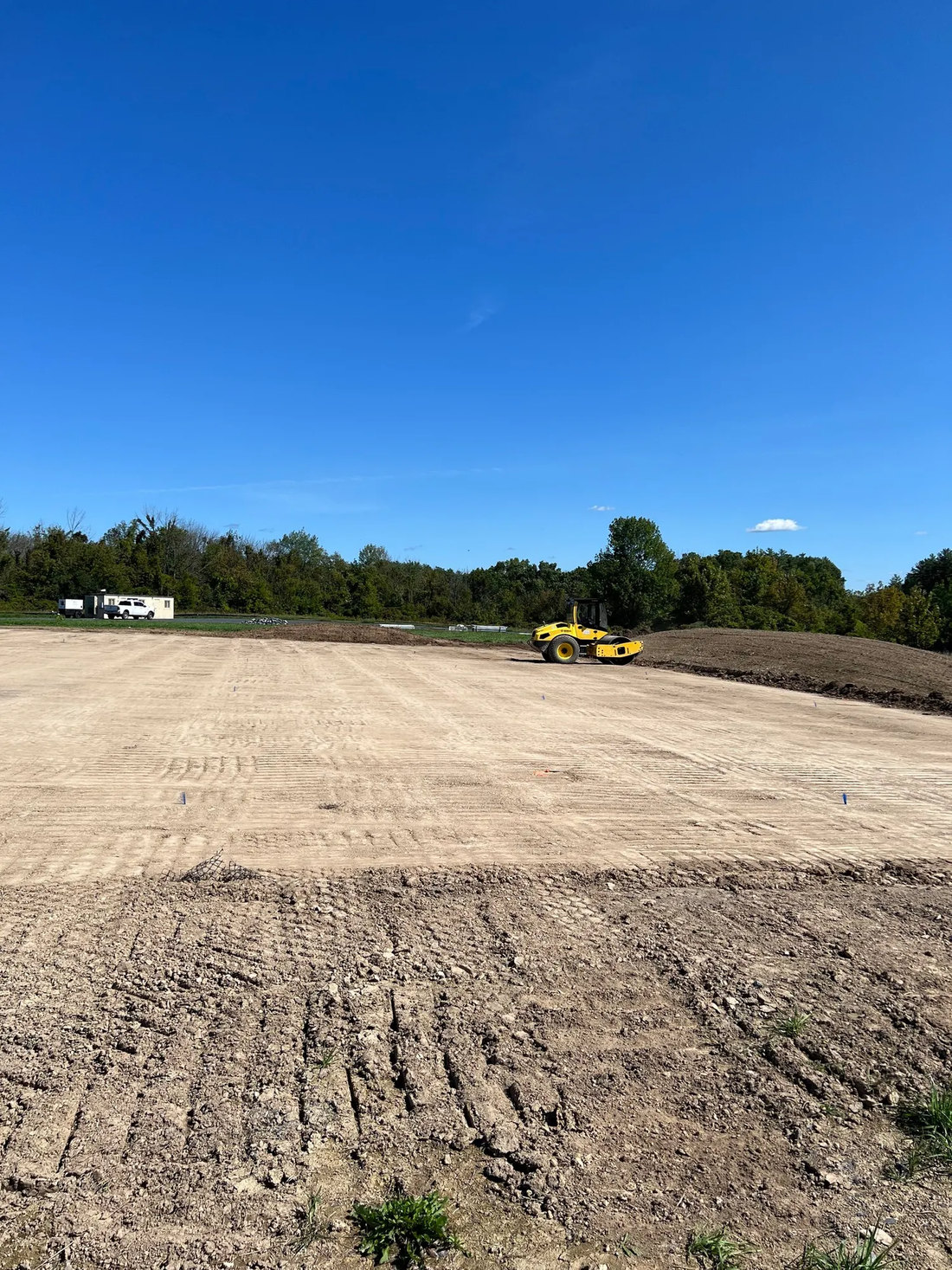 Construction site with leveled ground, heavy machinery, and trees under a blue sky.