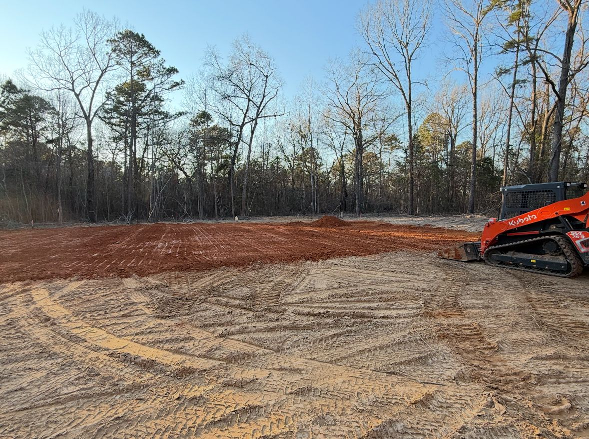 A skid steer clears red dirt on a construction site, with trees and blue sky in the background.